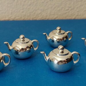 May include: Six small, shiny silver teapots are arranged on a blue surface. Each teapot has a rounded body, a curved spout, a small handle, and a lid with a knob. The teapots reflect the surrounding light.