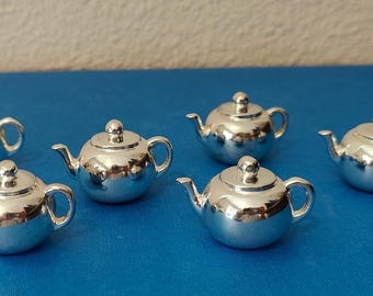 May include: Six small, shiny silver teapots are arranged on a blue surface. Each teapot has a rounded body, a curved spout, a small handle, and a lid with a knob. The teapots reflect the surrounding light.