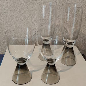 May include: Four clear wine glasses with gray-tinted bases. The glasses have a unique design, with a wide bowl and a stem that flares out at the base. The glasses are arranged on a white surface, with a book titled "Bedrooms" visible.
