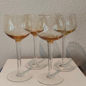 May include: Four clear wine glasses with amber-colored bowls and long stems. The glasses are arranged on a stack of books with titles visible, including "Bedrooms" and "Courtyard Gardens". The background is a textured, off-white wall.