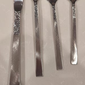 May include: Close-up of a set of silverware, including a knife, two forks, and a spoon. The silverware is silver-colored with a floral design on the handles. The utensils are arranged on a light-colored surface.