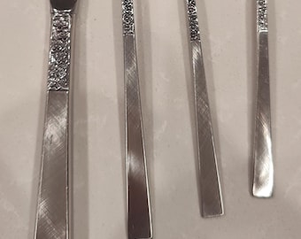May include: Close-up of a set of silverware, including a knife, two forks, and a spoon. The silverware is silver-colored with a floral design on the handles. The utensils are arranged on a light-colored surface.