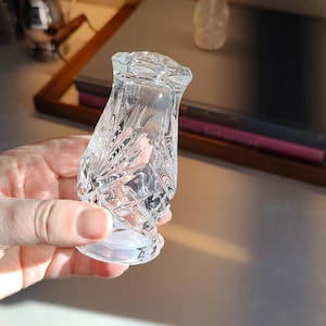 May include: Clear, cut-glass salt shaker with a diamond and fan pattern. The shaker has a flared base and a faceted top. A second, smaller shaker is visible in the background. The objects are on a light-colored surface.