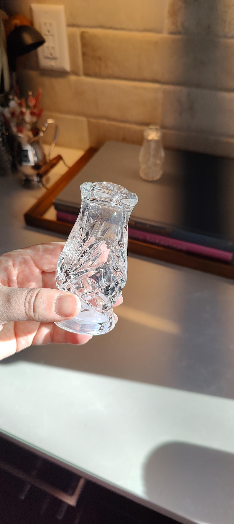 May include: Clear, cut-glass salt shaker with a diamond and fan pattern. The shaker has a flared base and a faceted top. A second, smaller shaker is visible in the background. The objects are on a light-colored surface.