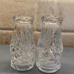 May include: Two clear, cut-glass salt and pepper shakers. The shakers have a diamond and fan pattern. The shakers are on a gray surface with a brick background.