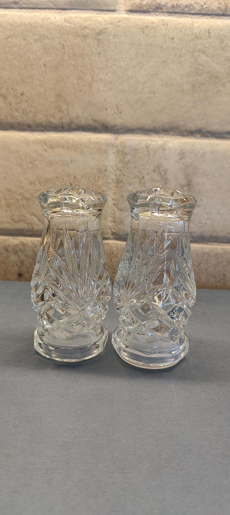 May include: Two clear, cut-glass salt and pepper shakers. The shakers have a diamond and fan pattern. The shakers are on a gray surface with a brick background.