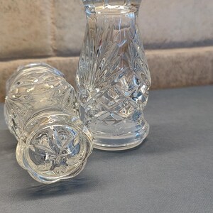 May include: Two clear, cut-glass salt and pepper shakers. One is upright, the other is on its side. The shakers have a diamond pattern and a decorative top. The background is a light gray brick wall.