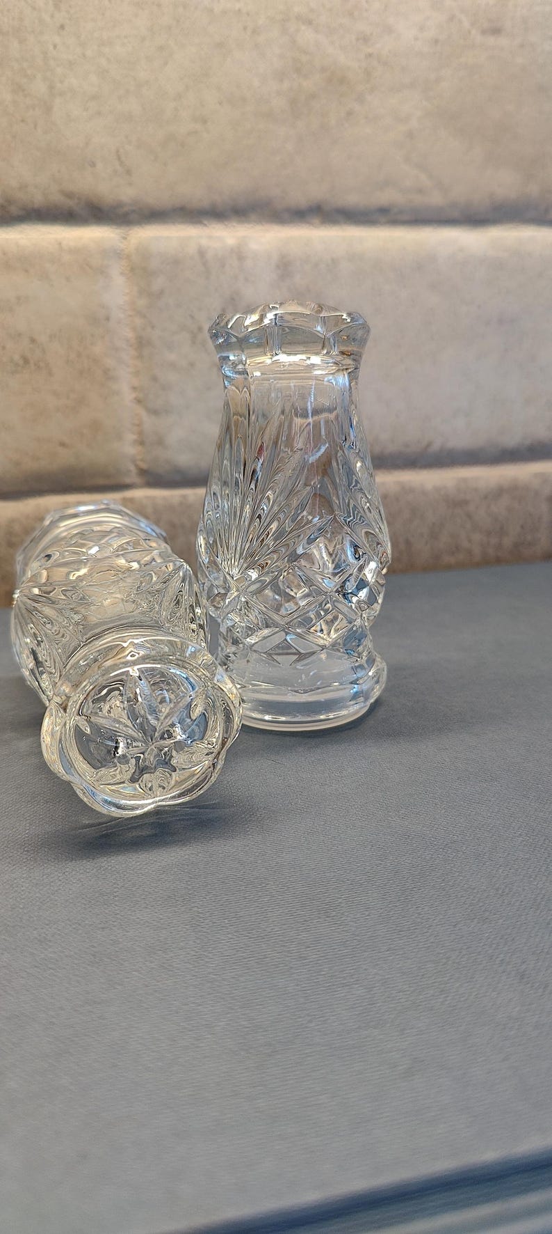 May include: Two clear, cut-glass salt and pepper shakers. One is upright, the other is on its side. The shakers have a diamond pattern and a decorative top. The background is a light gray brick wall.