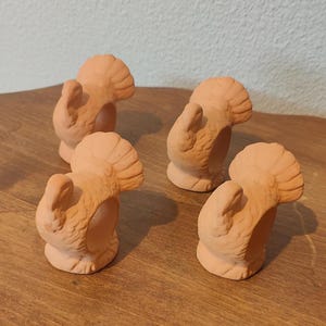 May include: A set of four terracotta-colored napkin rings, each shaped like a turkey with a fanned tail and textured plumage. The circular openings are designed to hold napkins, and the figures are rendered in a matte, earthy finish.