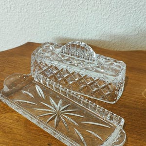 May include: A clear glass butter dish consisting of a rectangular base and a matching lid. The base features a starburst pattern in the center, while the lid has a diamond-cut geometric design and a curved handle on top for easy lifting.