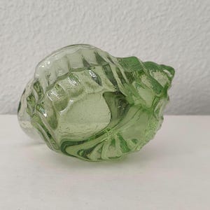 May include: A translucent light green glass seashell figurine featuring a molded spiral design with raised ridges and a smooth, glossy finish. The object has a hollow center and a curved, organic shape that mimics the appearance of a natural conch shell.