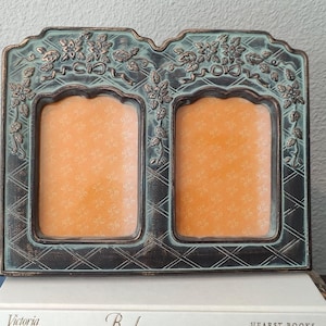May include: A decorative double photo frame with an ornate, aged teal and bronze finish. The frame features floral and ribbon detailing at the top and a diamond pattern on the sides. The two photo openings have an orange background.