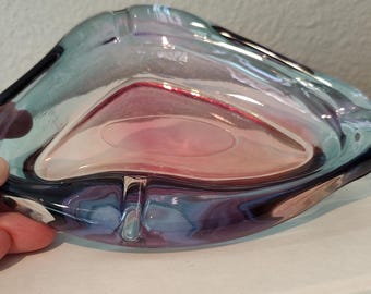 Könnte beinhalten: Ein mehrfarbiger Glasaschenbecher mit einer einzigartigen, organischen Form. Der Aschenbecher weist eine Mischung aus Blau-, Lila- und Rosatönen auf, die ein ansprechendes Design ergeben. Der Aschenbecher liegt auf einer weißen Oberfläche.