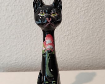 May include: A black ceramic cat figurine with green eyes, a pink nose, and a floral design on its neck. The cat has colorful socks and is sitting upright. The figurine is on a white surface.