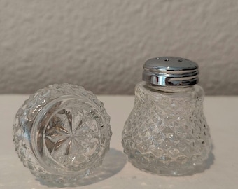May include: Two clear glass salt and pepper shakers with a diamond pattern. One shaker has a silver lid with small holes. The other shaker is upside down, revealing a starburst pattern on the bottom. The shakers are on a white surface.