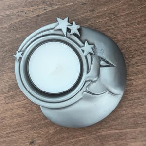 May include: A silver-toned candle holder shaped like a crescent moon with a face, featuring three small star accents along the upper edge. A round white tea light candle is seated securely within the circular center of the moon-shaped base.