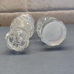May include: Two clear, cut-glass vases with a textured, diamond-like pattern. One vase is upright, and the other is lying on its side, revealing a white plastic base. The vases are on a blue surface.