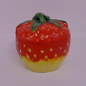 Può includere: Un contenitore in ceramica a forma di fragola con una glassa rossa e gialla. Il contenitore ha uno stelo e foglie verdi in cima.