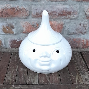 Può includere: Un barattolo per l'aglio in ceramica bianca a forma di viso sorridente con due fori nella parte superiore. Il barattolo è su un tavolo di legno.