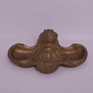 May include: A vintage brass inkwell with a decorative floral design. The inkwell has two shallow wells on either side and a central, lidded compartment. The brass has a warm, aged patina, suggesting its antique nature. The overall shape is symmetrical and ornate.