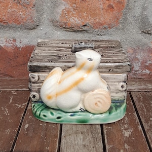 May include: A ceramic figurine of a white and orange rabbit sitting on a green base with a log-cabin-style box behind it. The figurine is set on a wooden surface with a brick wall in the background.