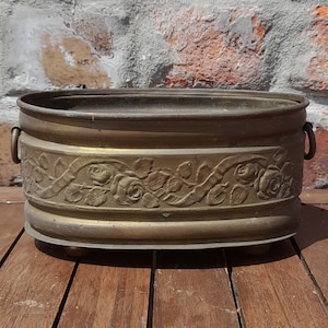 May include: An oval brass planter with a decorative floral design and two handles. The planter is sitting on a wooden surface.