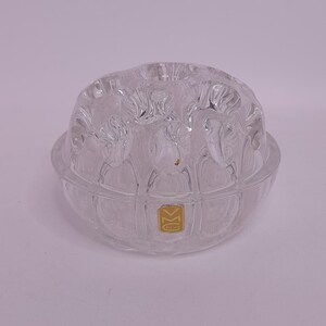 May include: A clear glass bowl with a textured, rounded top and a shallow base. The top has a floral or leaf-like pattern. A small, gold-colored sticker is visible on the base. The bowl is likely for decorative or functional use.