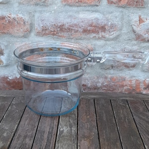 May include: A clear glass bowl with a silver rim and a handle. The bowl has a light blue accent line and sits on a weathered wooden table. The background is a brick wall.