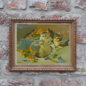 Op de afbeelding: Een ingelijste vintage prent met katten. Het kunstwerk toont katten bij een vissenkom en boeken, met een warm kleurenpalet. De lijst is sierlijk en goudkleurig, wat bijdraagt aan de antieke uitstraling. De achtergrond is een verweerde bakstenen muur.
