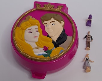 Könnte beinhalten: Ein rosa Plastik-Kompakt mit einem Bild einer Prinzessin und eines Prinzen im Inneren. Das Kompakt kommt mit drei kleinen Plastikfiguren einer Prinzessin, eines Prinzen und einer Fee.