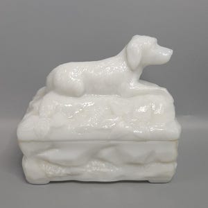 Könnte beinhalten: Eine weiße Keramik-Schmuckschatulle mit einer Hundefigur oben. Der Hund liegt und die Schatulle hat ein strukturiertes, felsiges Aussehen. Es ist ein dekorativer Gegenstand.