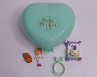 May include: A teal heart-shaped Polly Pocket case with a gold Polly Pocket logo. The case is open and there are several small plastic toys inside, including a doll, a bed, and a ring.