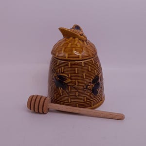 Può includere: Un vasetto di miele in ceramica marrone con coperchio, con un design a cestino intrecciato e illustrazioni di api. Un cucchiaio di miele in legno è appoggiato accanto al vasetto. Un affascinante accessorio da cucina.