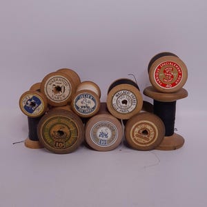 Könnte beinhalten: Eine Sammlung von Vintage-Holzspulen mit Faden, jede mit einem anderen Etikettendesign. Die Spulen variieren in Größe und Farbe, einige mit schwarzem Faden und andere ohne sichtbaren Faden. Die Etiketten zeigen Text und Grafiken.