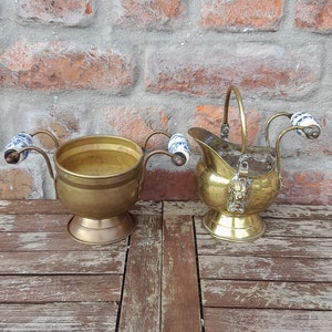 Könnte beinhalten: Zwei antike Messingkrüge mit Porzellanhenkeln. Die Krüge stehen auf einem Holztisch vor einer Backsteinwand.