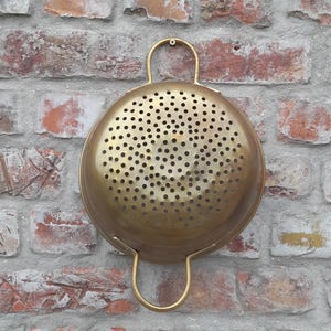 Può includere: Una scolapasta rotonda color ottone con numerosi piccoli fori e due manici curvi. La scolapasta è montata su un muro di mattoni usurati, che ne evidenzia l'estetica vintage. Il muro di mattoni presenta un mix di mattoni rossi, marroni e grigi.