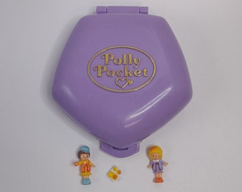 Puede incluir: Un juguete Polly Pocket morado con un logotipo dorado y dos pequeñas figuras. Las figuras son una niña con cabello rubio y un niño con cabello castaño.