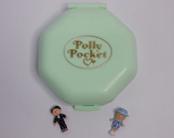 Könnte beinhalten: Ein hellgrünes Polly Pocket Spielzeug mit einer goldenen Aufschrift "Polly Pocket". Zwei kleine Plastikfiguren, eine in einem blauen Anzug und eine in einem blauen Kleid, stehen vor dem Spielzeug.