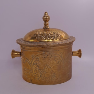 Könnte beinhalten: Ein goldfarbener Metallbehälter mit Deckel. Der Behälter hat zwei Griffe und ist mit floralen Mustern verziert. Der Deckel ist mit einem dekorativen Knauf versehen. Der Behälter wird wahrscheinlich zur Aufbewahrung oder Präsentation verwendet.