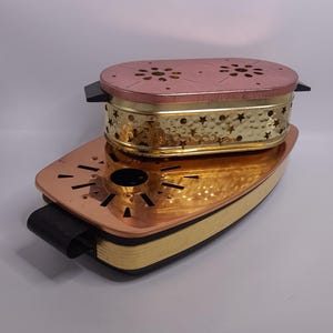 Könnte beinhalten: Zweiteiliges Räuchergefäß-Set aus Metall. Die Basis ist kupferfarben mit einem schwarzen Griff, und das Oberteil ist goldfarben mit einem perforierten Sternmuster. Der Deckel ist roségoldfarben mit dekorativen Aussparungen.