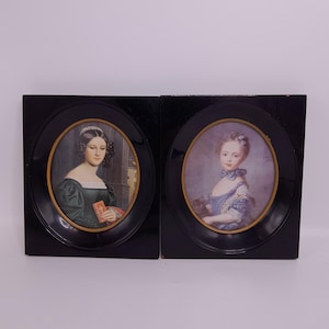 Può includere: Due miniature di ritratti antichi in cornici ovali nere. I ritratti raffigurano donne in abiti d'epoca, una in un abito verde con un libro, l'altra in un abito blu. Le cornici hanno dettagli dorati.