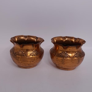 Könnte beinhalten: Zwei kleine, kupferfarbene dekorative Töpfe mit strukturierter Oberfläche. Jeder Topf hat einen gerüschten Rand und einen abgerundeten Körper mit einem geprägten Blumendesign. Die Töpfe werden vor einem schlichten weißen Hintergrund präsentiert.