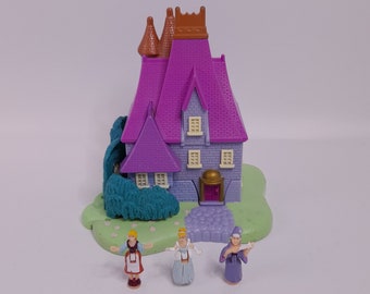 May include: A purple plastic playhouse with a green bush and three small figures in front of it. The figures are a woman in a red dress, a woman in a white dress, and a woman in a purple dress.