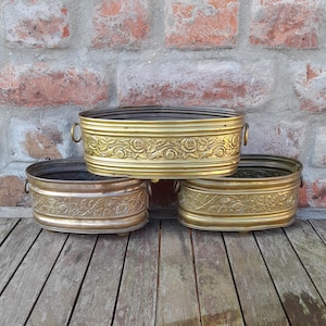 Op de afbeelding: Drie ovale plantenbakken van antiek messing met bloemmotieven. De plantenbakken, van verschillende afmetingen, staan op een verweerd houten oppervlak tegen een bakstenen muur. Elk heeft kleine zijhandvatten.