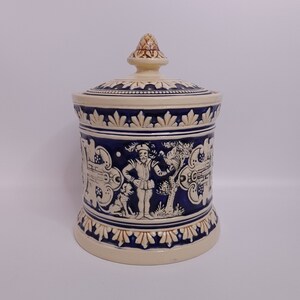 以下が含まれることがあります： 青と茶色のデザインが施された白い陶器の壺。壺には尖った蓋が付いています。壺には、犬と木と一緒にいる男性のシーンが描かれています。