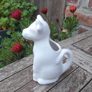 May include: A white ceramic cow-shaped jug with a handle. The jug is on a weathered wooden table. Red flowers and green plants are in the background.