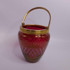 Op de afbeelding: Een robijnrode glazen vaas met een goudkleurige metalen handgreep en rand. De vaas heeft een ruitpatroon dat in het onderste gedeelte is geëtst. De handgreep is gebogen en de rand heeft een decoratief ontwerp.