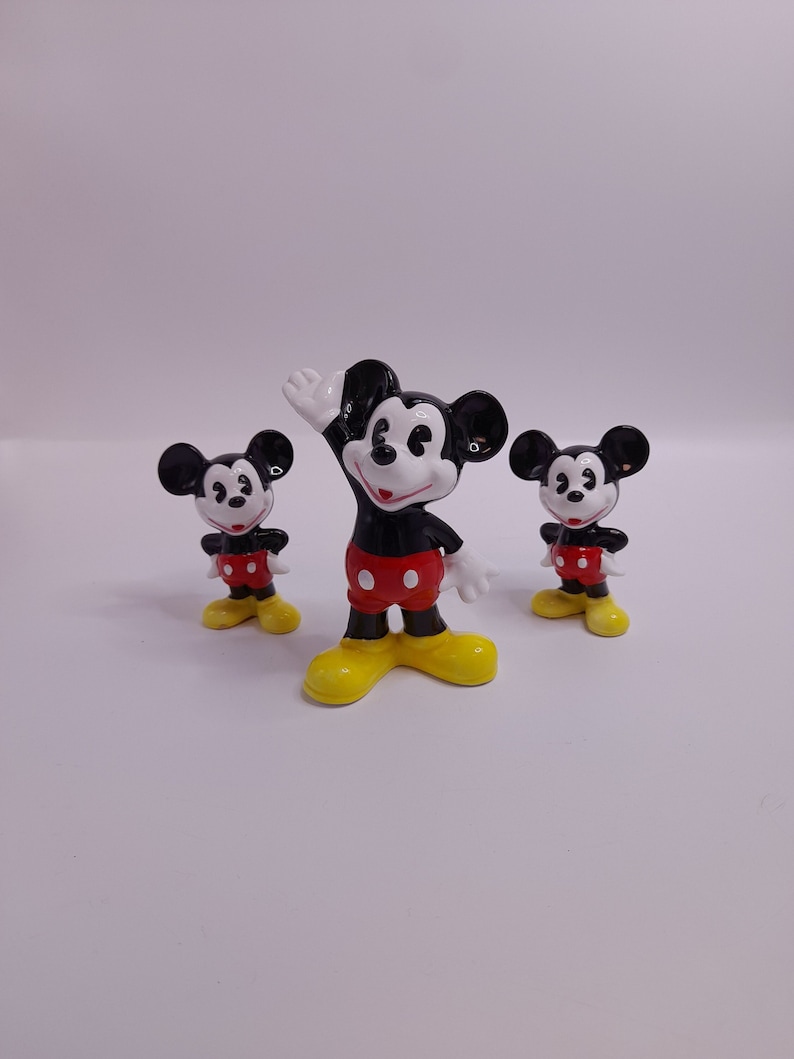 May include: Three ceramic Mickey Mouse figurines in different sizes. The largest figurine is standing with its right arm raised, while the two smaller figurines are standing with their arms at their sides. All three figurines are black with white eyes and red shorts with black buttons.