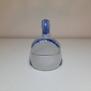 May include: A small, white ceramic trinket box shaped like a duck. The duck is painted in shades of blue and white.