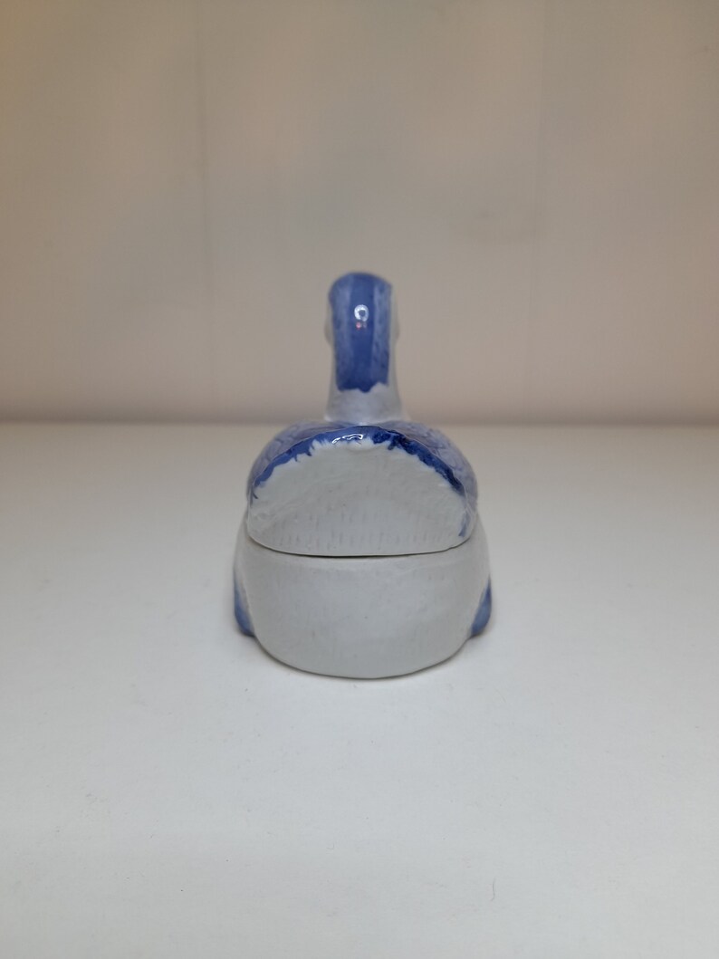 May include: A small, white ceramic trinket box shaped like a duck. The duck is painted in shades of blue and white.